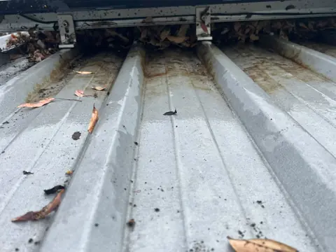 Dry leaves packed into the gap under the edge of a solar panel between the mounting feet, with rust stains in the channels of the metal roof