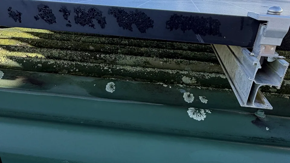 The open gap under the edge of a rooftop solar panel, with lichen on the metal roof below