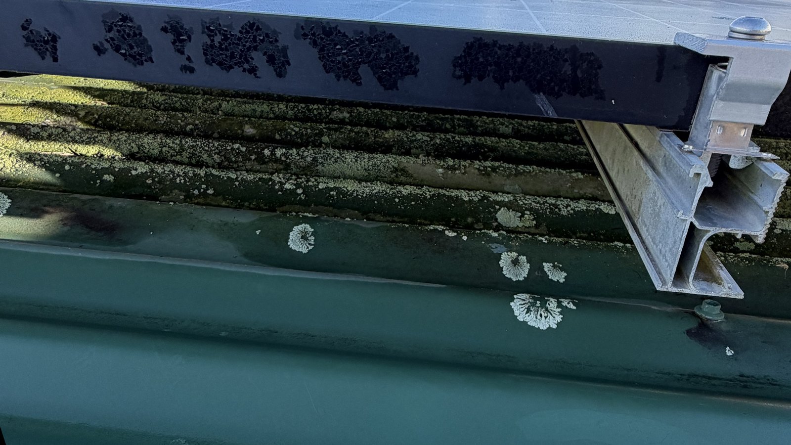 The open gap under the edge of a rooftop solar panel, with lichen on the metal roof below